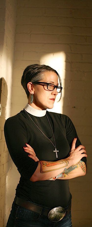 Nadia Bolz-Weber posing with her arms crossed and wearing a black pastors outfit, jean pants and a rosary.