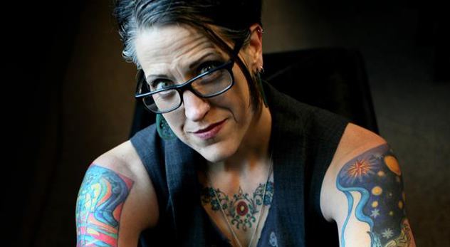 Nadia Bolz-Weber sitting down with her tattoos visible and wearing a black sleeveless shirt and eyeglasses.