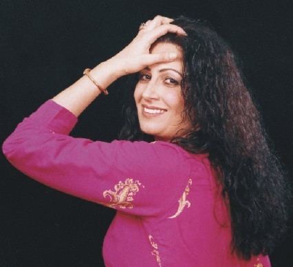 Naghma smiling in a side pose, with her left hand touching her forehead, has a curly black hair, she has a gold ring and bracelet, and wearing a pink 3/4-length sleeve top