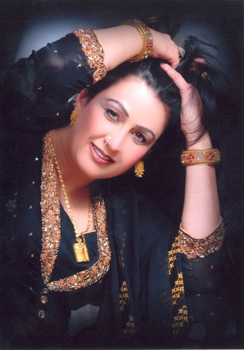 Naghma smiling with both hands lifting her hair upward, she has a makeup, black hair, a piercing on her nose, a gold necklace, bracelets, gold dangling earrings, and a ring wearing a black and gold salwar kurta
