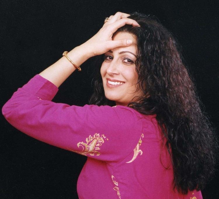 Naghma smiling in a side pose, with her left hand touching her forehead, has a curly black hair, has a gold ring and bracelet, and wearing a pink 3/4-length sleeve top