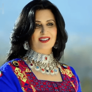 Naghma smiling looking at the ground, she has a makeup, black hair, a piercing on her nose, wearing a choker, silver round earrings and a colored blouse