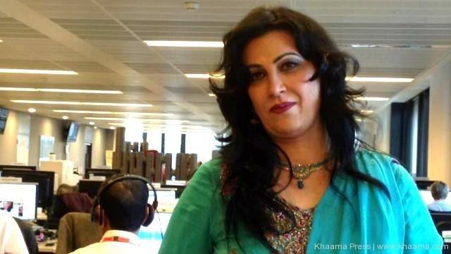 Naghma smiling, she has a light makeup, a black curly hair, and a blue necklace inside a building with people in the background facing their computers, wearing a blue salwar kurta