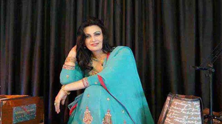 Naghma smiling with her right hand touching her hair and left arm resting on her knee while sitting, she has a makeup, a black hair, a gold bracelet, a necklace, and a ring, with a black curtain in the background with a bag on her right and a Harmonium instrument on her left, she's wearing a blue salwar kurta