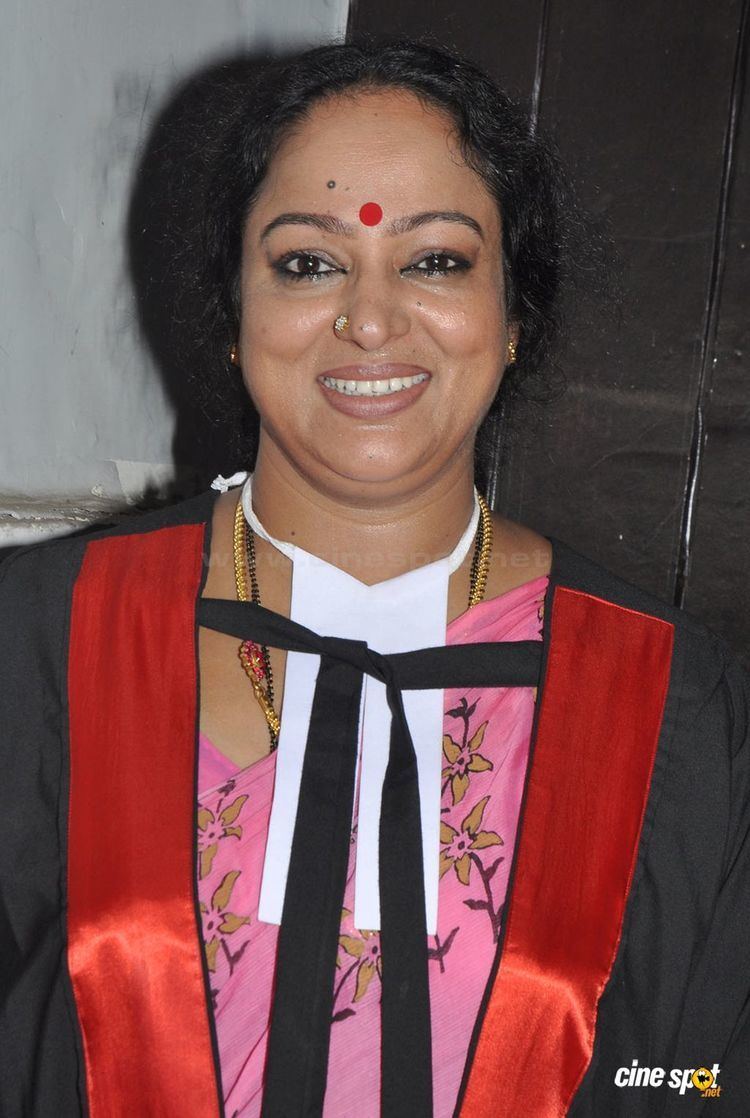 Nalini smiling while wearing a black and red blazer, pink floral blouse, earrings, and necklace