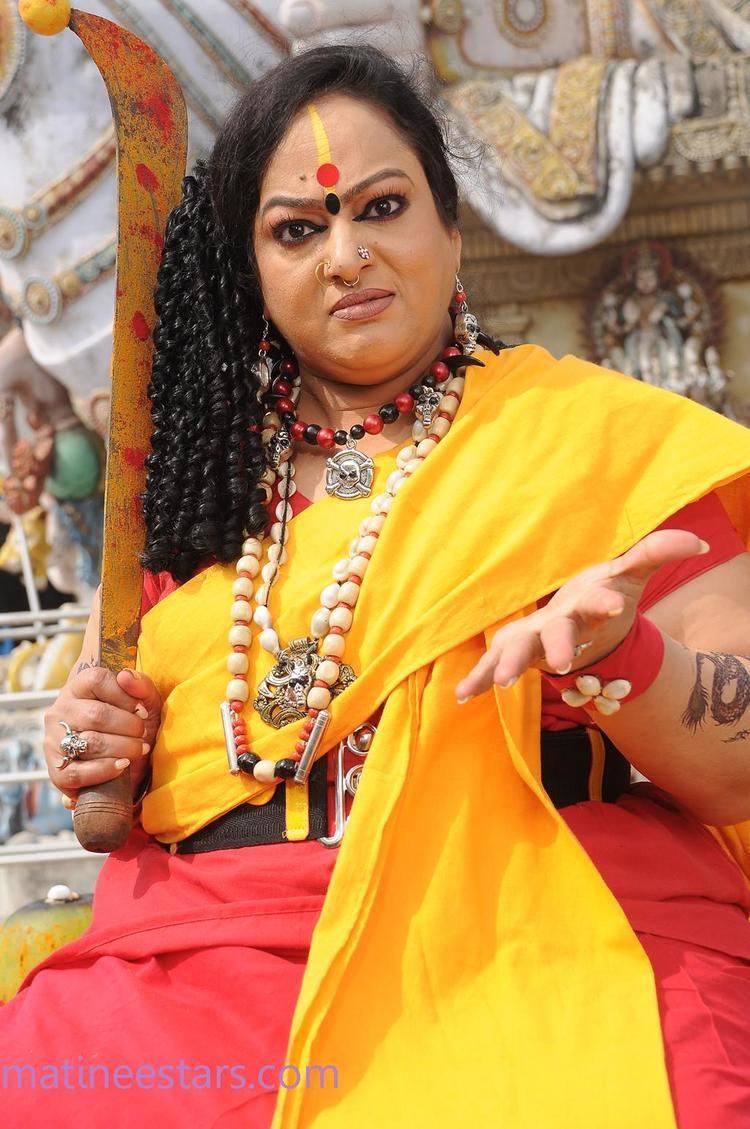 Nalini with an angry face and curly hair while wearing a yellow and orange dress, nose jewel, skull ring, skull earrings, and necklaces