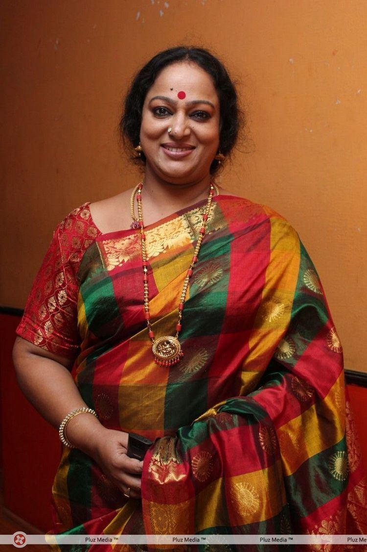 Nalini smiling while wearing a red, yellow, and green checkered dress, gold earrings, necklaces, and bracelet