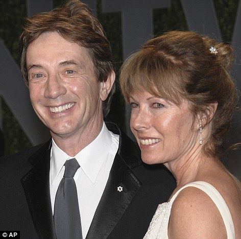 Nancy Dolman and Martin Short are smiling. Nancy with blonde hair, wearing earrings, and a white sleeveless dress while Martin wearing a black coat over white long sleeves, and a gray tie.