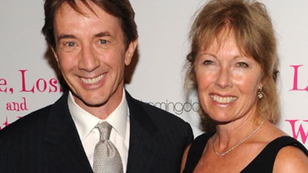 Nancy Dolman and Martin Short are smiling. Nancy with blonde hair, wearing earrings, a necklace, and a black sleeveless top while Martin wearing a black coat over white long sleeves, and a gray tie.