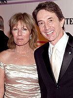 Nancy Dolman with a serious face while Martin Short smiling. Nancy with blonde hair and wearing a gold off-shoulder dress while Martin wearing a black coat over white long sleeves, and a gold tie.
