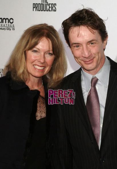 Nancy Dolman and Martin Short are smiling. Nancy with blonde hair, wearing a necklace, and a black blazer over a black lace top while Martin wearing a black striped coat over gray long sleeves, and a brown tie.