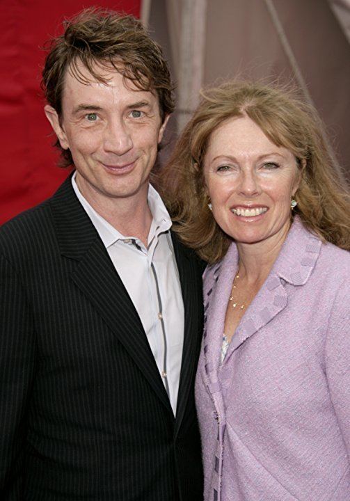 Nancy Dolman and Martin Short are smiling. Nancy with blonde hair, wearing earrings, a necklace, and a purple blazer while Martin wearing a black striped coat over white long sleeves.