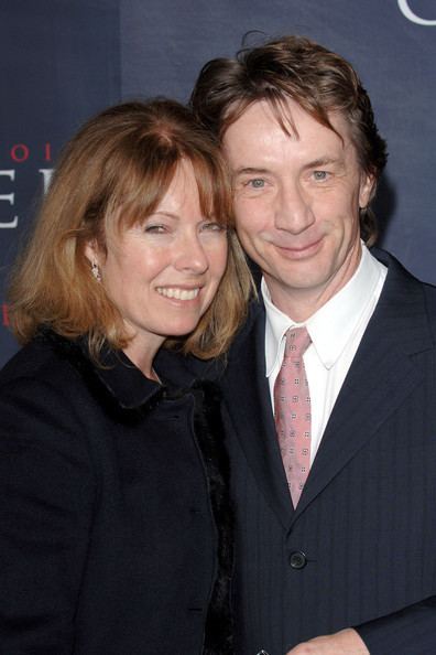 Nancy Dolman and Martin Short are smiling. Nancy with blonde hair, wearing earrings, and a black coat while Martin wearing a black striped coat over white long sleeves, and a brown tie.