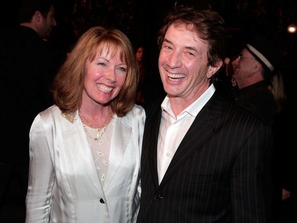 Nancy Dolman and Martin Short are smiling. Nancy with blonde hair, wearing a necklace, and a white coat over a white top while Martin wearing a black striped coat over white long sleeves.