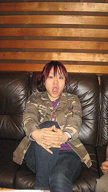 Nao Kawakita’s mouth opened, sitting down on a black couch has light violet hair with bangs, both hands on her left leg lifting it up, wearing a  white wristwatch on her right hand, a violet t-shirt, and a military jacket.