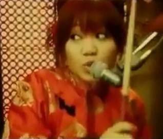 Nao Kawakita is serious, both eyes looking at her right, mouth half opened, has a blend of red and brown hair with bangs, her left hand holding a drum stick, microphone in front, wearing a red kimono with a black and gold design.