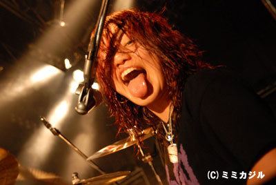 Nao Kawakita sticks out her tongue, has a blend of red and brown curly hair, a microphone in front, and a cymbal drum stand (left), wearing a silver necklace and black t-shirt with a minimalistic violet design.