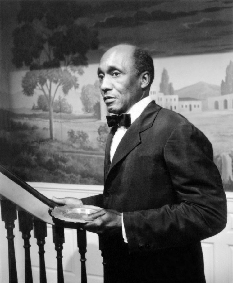 Napoleon Whiting holding a plate while standing beside a staircase (black and white)