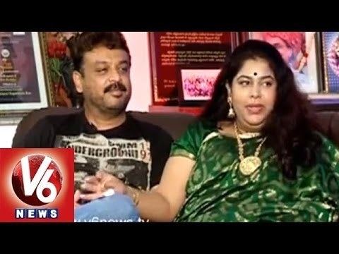 Naresh smiling with a mustache and wearing a black shirt with his wife sitting beside him wearing a green saree, gold necklace and dangling earrings