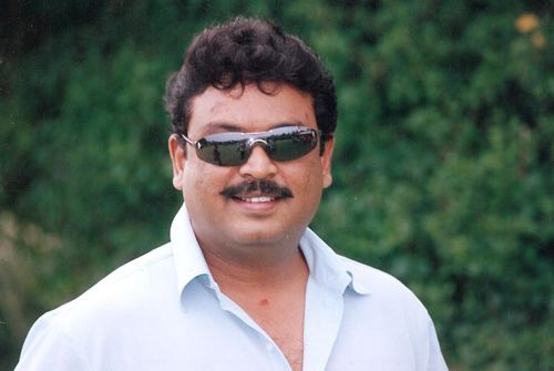 Naresh smiling with curly hair and mustache while wearing sunglasses and a white shirt