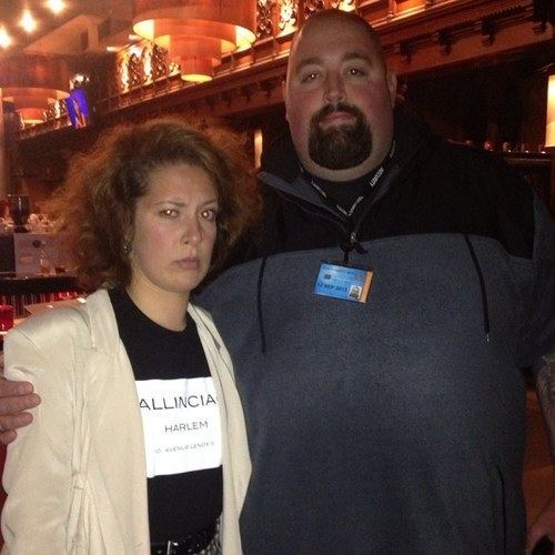 Natalie Casey with a strict face and curly hair, wearing a blazer and a black shirt together with a man with a beard and wearing a black polo shirt.