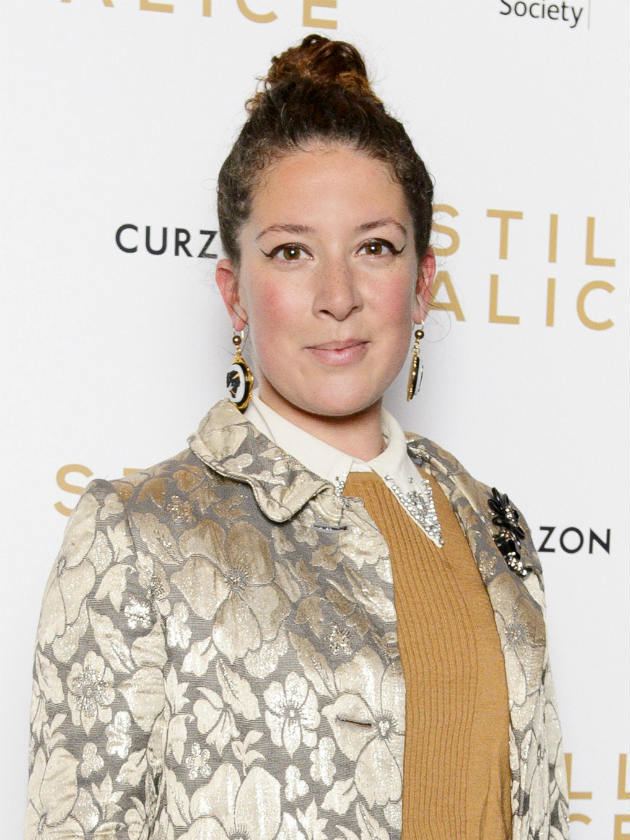 Natalie Casey smiling and wearing earrings, a floral blazer, and a yellow blouse.