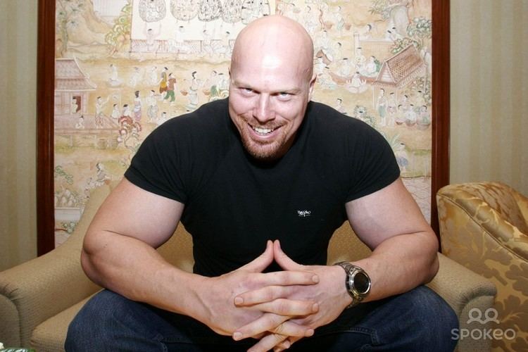 Nathan Jones, smiling with a ferocious look in his eyes, with crossed fingers and arms resting on top of his legs while sitting on a couch and a mural painting on his back, he has a bald head, a beard, and a mustache. He is wearing a black shirt, jeans, and a watch on his wrist