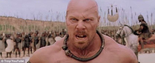 Nathan Jones as Boagrius, a scene from the 2004 movie "TROY. He looks angry facing someone and soldiers on his back, he has a bald head, a beard and mustache, and scars on his face, topless, and wearing a bronze necklace
