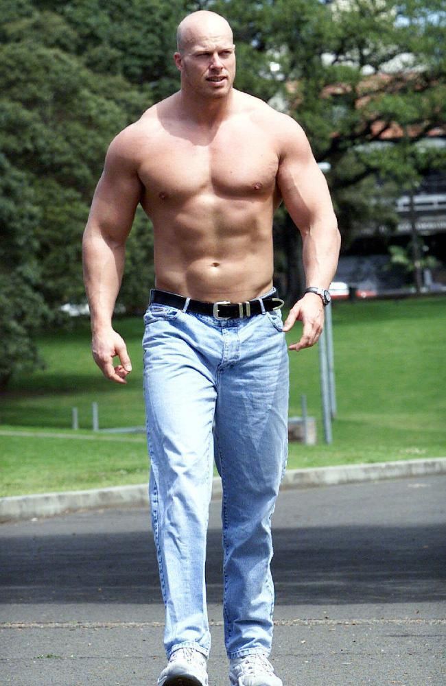 Nathan Jones, seriously looking afar while walking on the street and a tree in the background, has a bald head, 4 pack abs, topless, with a watch on his wrist, and wearing pants with a black belt and white shoes.