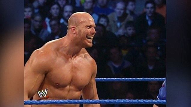 Nathan Jones shows an evil laugh inside the wrestling ring with an audience behind his back, has 4 pack abs, a bald head, and topless