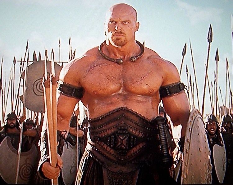 Nathan Jones seriously looking from afar, carrying spears and armor, with soldiers on his back in a scene from the 2004 movie "TROY", he has a bald head, a beard and mustache, and scars on his face and body, topless, wearing armlets, arm gloves, a bronze necklace, and an armor suit