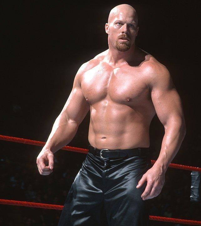 Nathan Jones seriously looking at someone inside a wrestling ring and an audience behind his back. He has 4 pack abs, a bald head, a beard & mustache, is topless, and wearing glossy black pants with a belt