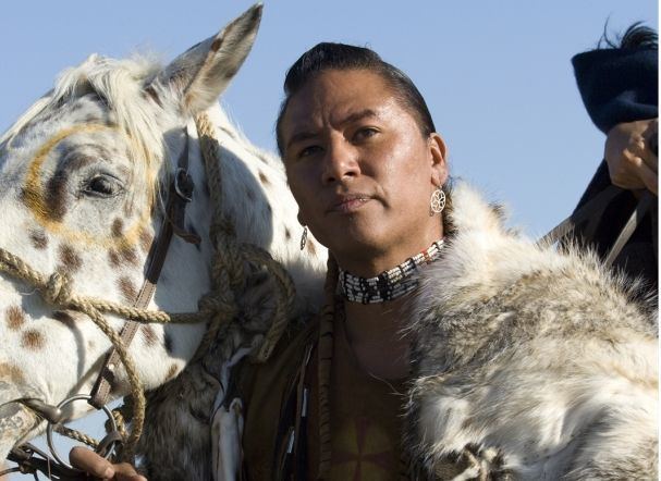 Nathan Lee Chasing His Horse wearing feather cloth, earrings, and necklace while beside him is a white speckled horse