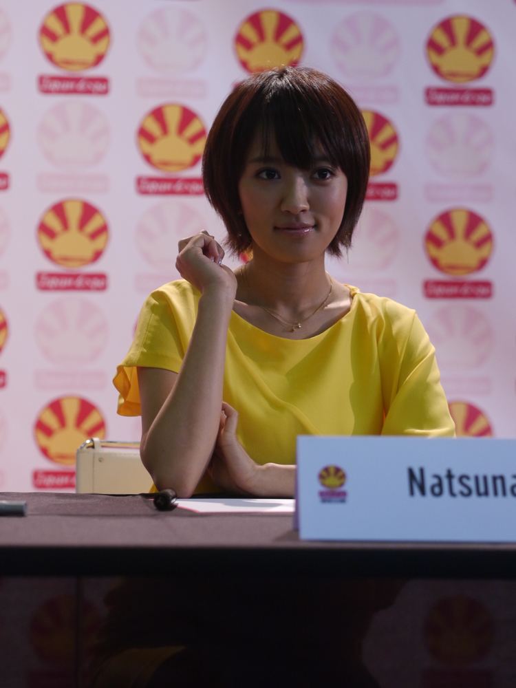At the Japanese Expo Festival, Natsuna Watanabe is smiling, and has hazelnut hair, both hands on the table, behind is the logo of the Japanese Expo, and she is seated on a white chair, in front is a table with her name, a paper, and pen, she is wearing a gold necklace and a yellow top.
