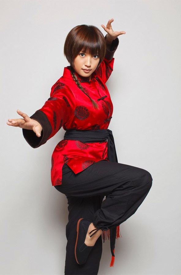 Natsuna Watanabe is serious, has chestnut braided hair, both hands up, right leg up, wearing a Japanese red costume on top with a black belt, black pants, and black-red shoes.