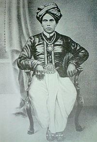 Tiruvarudi Vaihunda Nadan sitting on the chair while wearing a turban, long sleeves, pants, shoes, necklace, and rings
