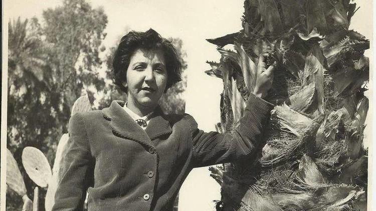 Naziha Selim seriously looking while holding a tree with her left hand and a cactus and trees in the background, a photograph in Baghdad in the early 1950s, she has short black hair, wearing a white polo with a little ribbon under a black coat