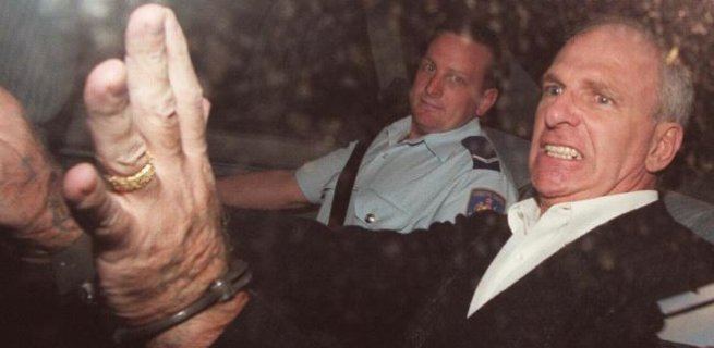 Neddy Smith sitting next to the police officer inside the car with an angry face while wearing a black coat and white long sleeve