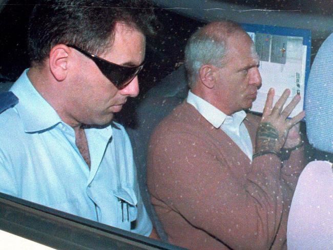 Neddy Smith covering his face while sitting next to the police officer  inside the car and wearing a brown blazer and white long sleeves