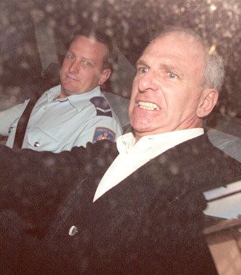 Neddy Smith sitting beside the police officer inside the car with an angry face while wearing a black coat and white long sleeve
