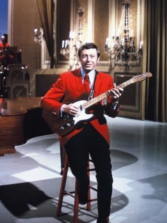 Neil Levang smiling while sitting on the chair and playing the guitar and wearing a red coat, white long sleeves, red necktie, and black pants