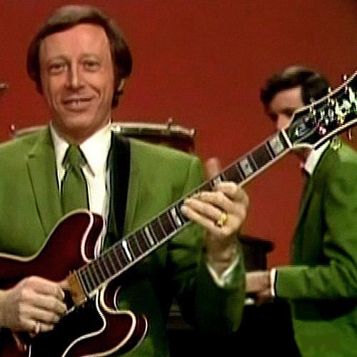 Neil Levang smiling while playing the guitar and wearing a green coat, white long sleeves, green necktie, and ring