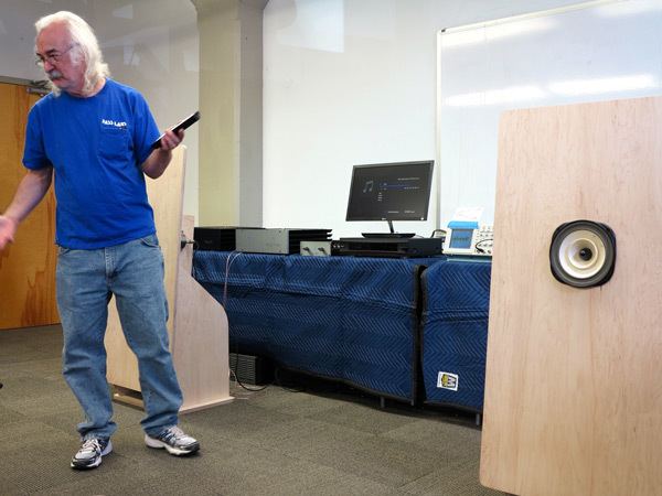 Nelson Pass standing and holding a remote in his left hand with a monitor on the table and a whiteboard on its back and on the right side is an audio amplifier. Nelson has white hair with a white beard and a mustache and wearing eyeglass, shoes, a blue shirt, and denim pants