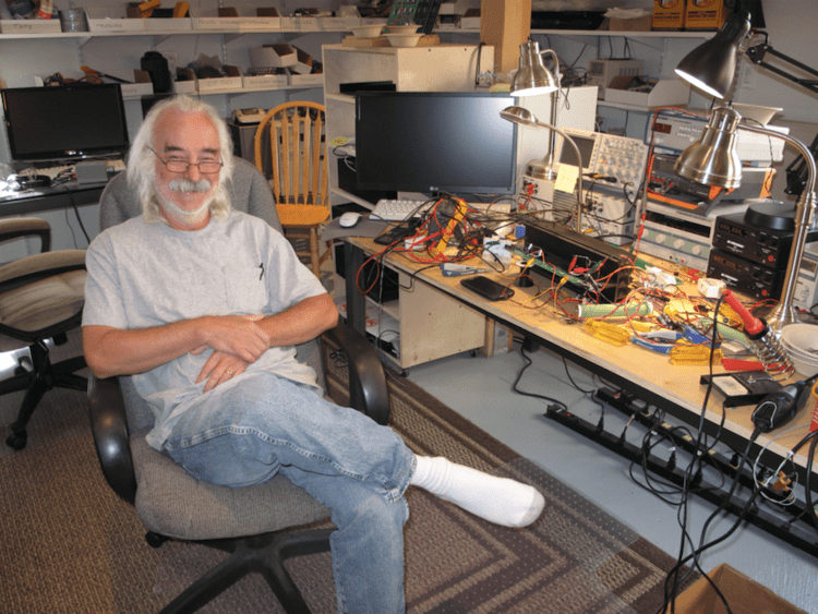 Nelson Pass smiling while sitting on an office chair and holding his left hand on his right hand with his left knees under his right feet in an office room with a monitor on the table, a wooden chair, an office chair, in the background, and beside him is a table with a monitor, 4 lamps, audio amplifiers, electric wires on it. Nelson has white hair with a white beard and mustache and wearing an eyeglass, white socks, gray shirts, and denim pants