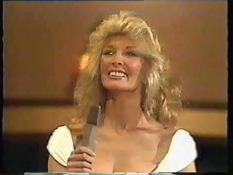 Marti Caine with a big smile, wavy blonde hair, wearing a white sexy top while holding a microphone.