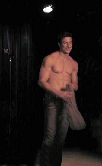 Nick Adams is smiling, has black hair, both hands holding a gray shirt inside a studio, on his right is a microphone, topless, wearing ash gray pants.
