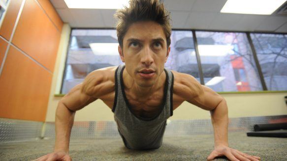 A man is serious, has black hair, both hands on the ground, doing push-ups inside a room, with a brown wall and glass window, wearing a dark gray with black lines jersey tank.