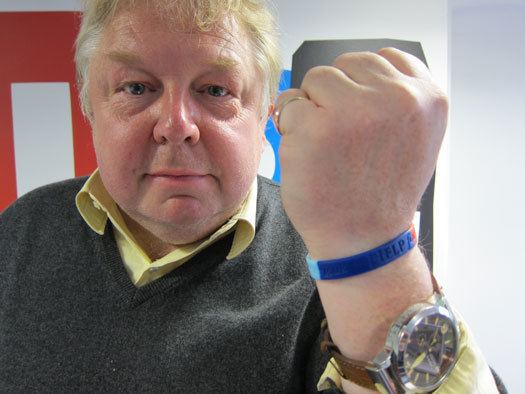 Nick Ferrari on a closed fist pose wearing a yellow and gray shirt, a watch, and a blue hand strap