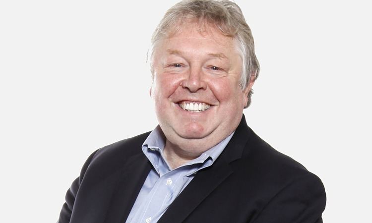 Nick Ferrari smiles while wearing a blue shirt and a black coat