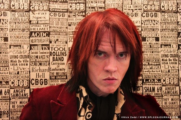 Nick Zedd with a serious face, long red hair, and wearing a red coat over an off-white and black long sleeve.